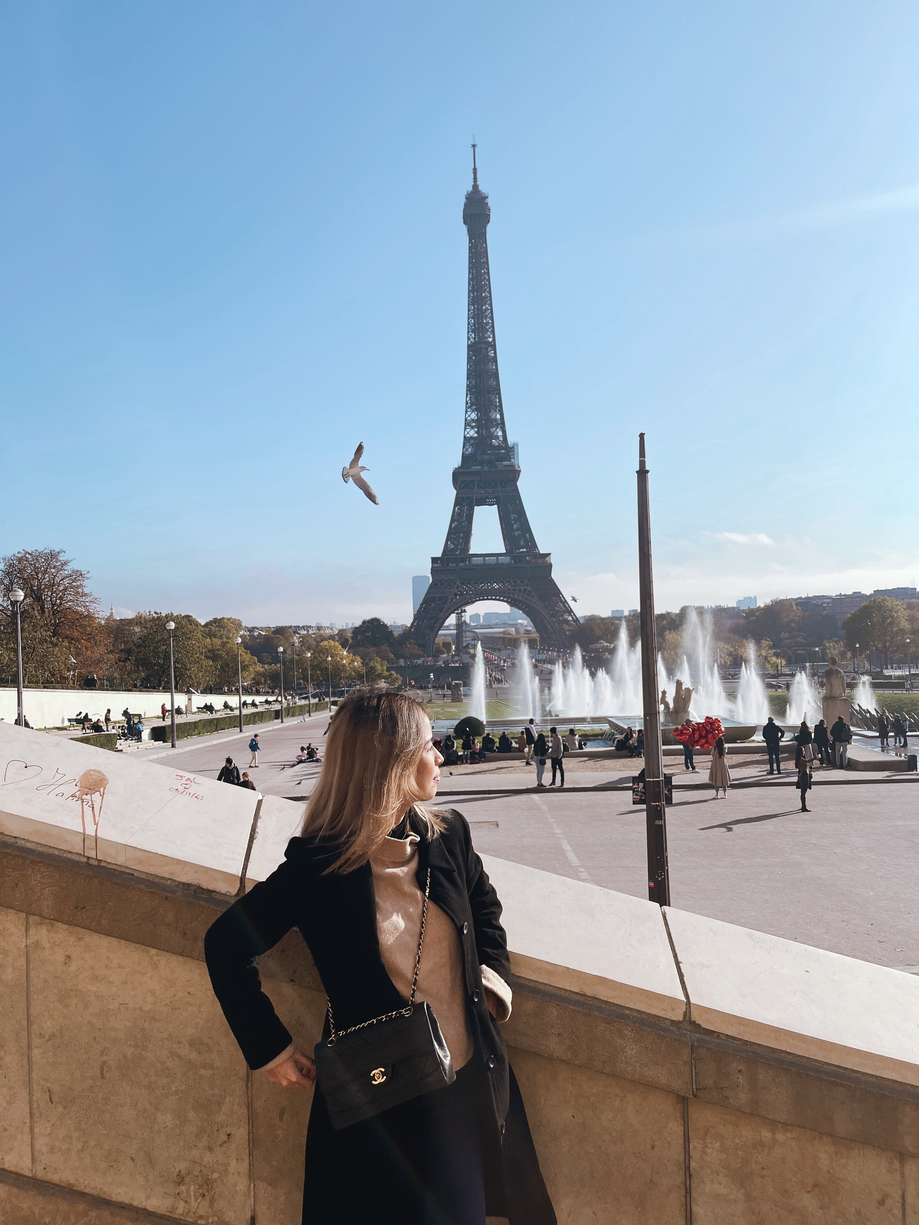 A woman in a black coat at the Trocadéro in Paris, with the Eiffel Tower and fountains behind her