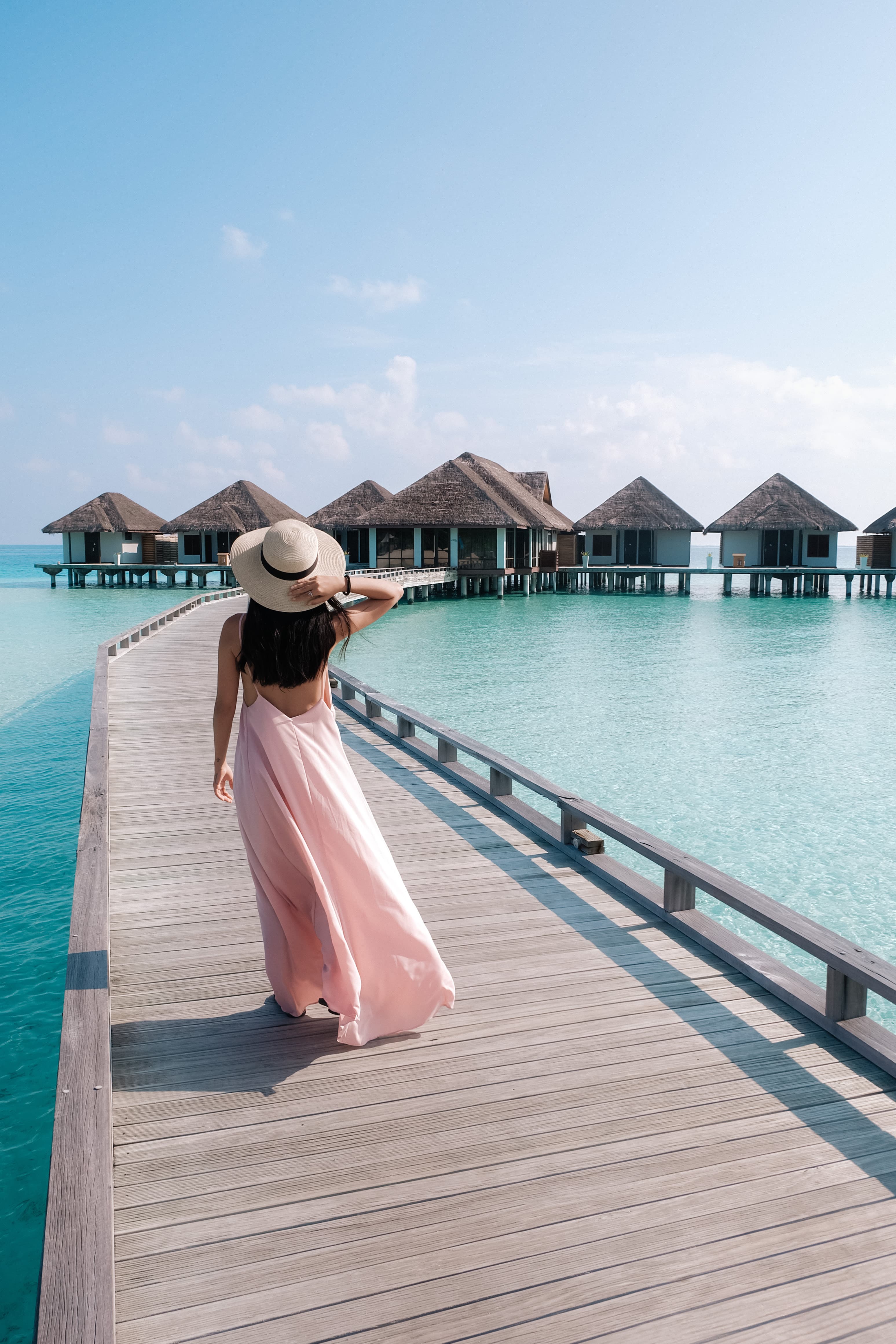 A woman in a pink dress and straw hat walking along a boardwalk towards overwater villas