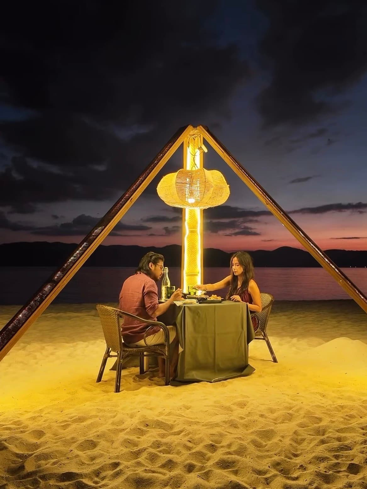 A couple having dinner on a beach at dusk under a glowing rattan lamp